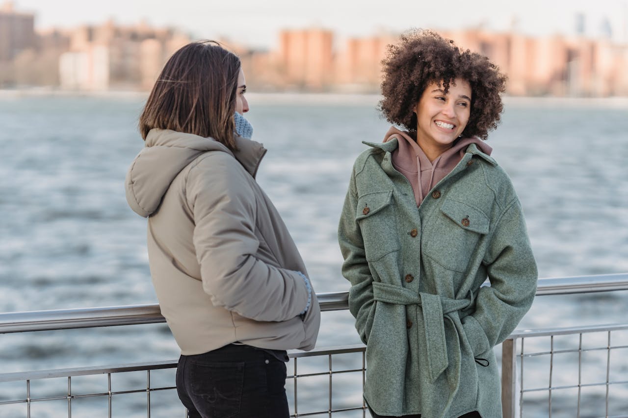 Services-01 Smiling diverse women in warm clothes standing near metal railing on embankment on background of river and opposite shore with houses in city
